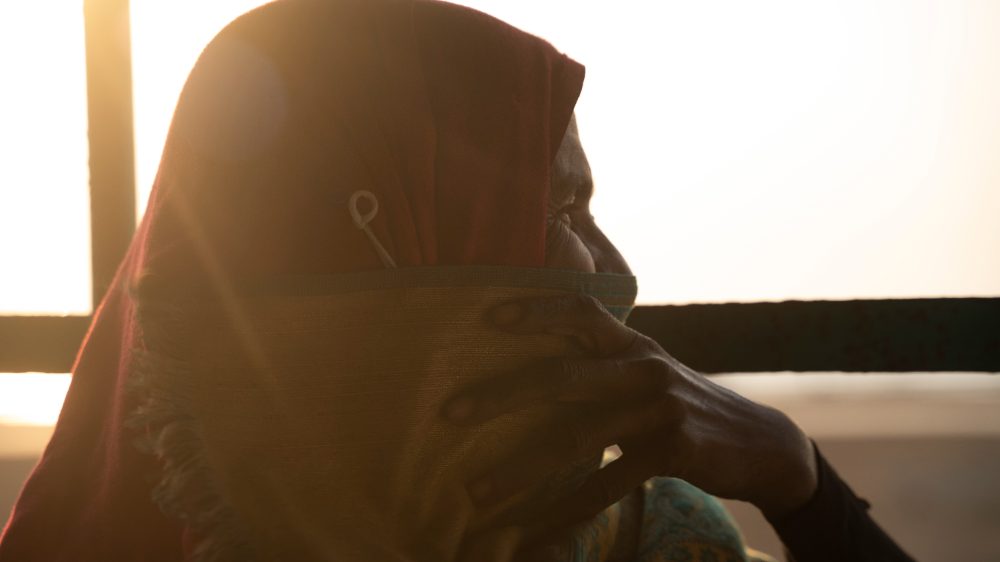 Amena in profile against low golden sunlight, her red headscarf drawn across her face, her hand at her cheek.