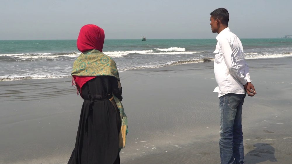 Amena, in a red headscarf, and Abdus stand on a grey beach, looking out to sea.